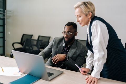 Middle-adult female Caucasian mentor supervisor help male african intern explain computer work corporate software. Businesswoman instruct trainee new worker teaching giving advice at workplace.